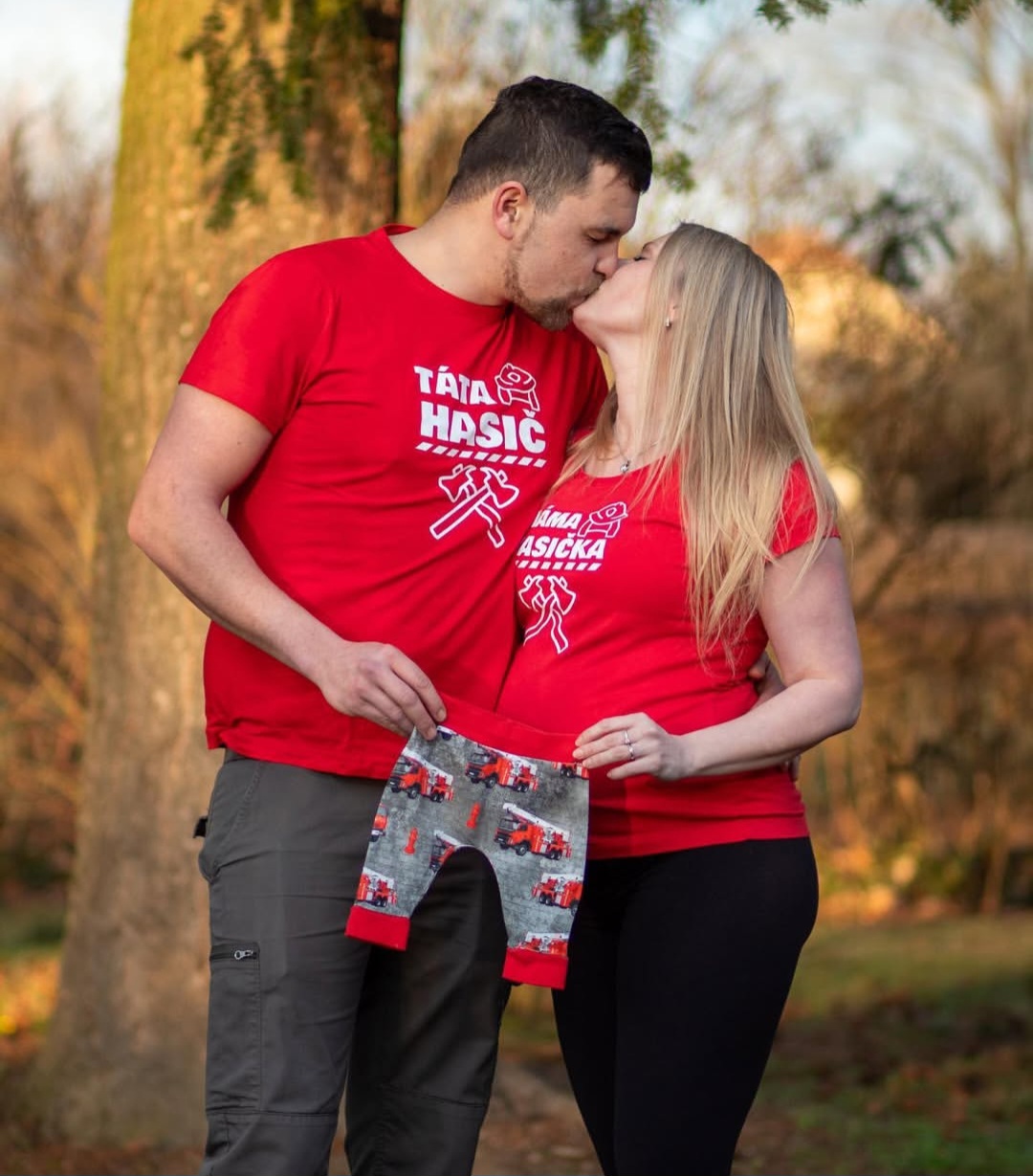 Těhotný pár v červených tričkách s dětským oblečením - oznamování pohlaví